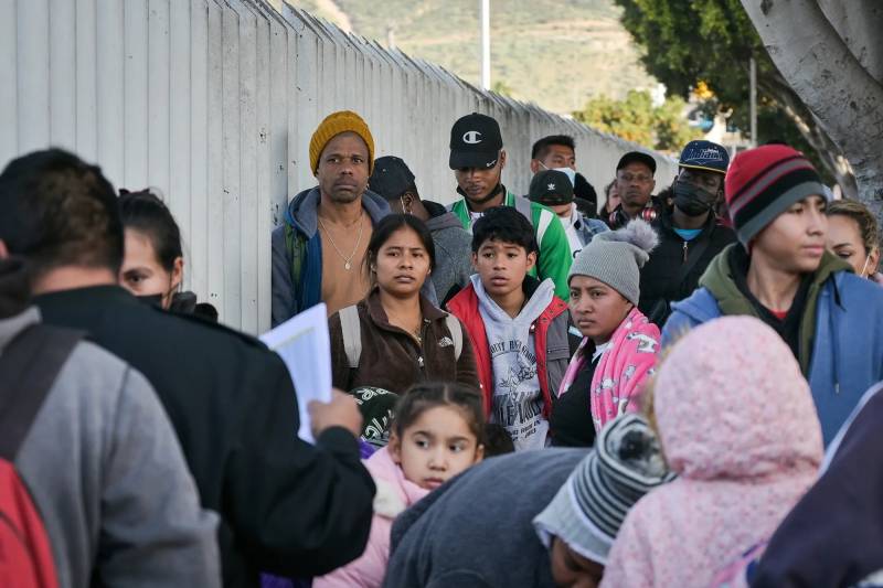 Latinx migrants of various ages from adults to children look on with concerned expressions as they stand next to white steel barriers.