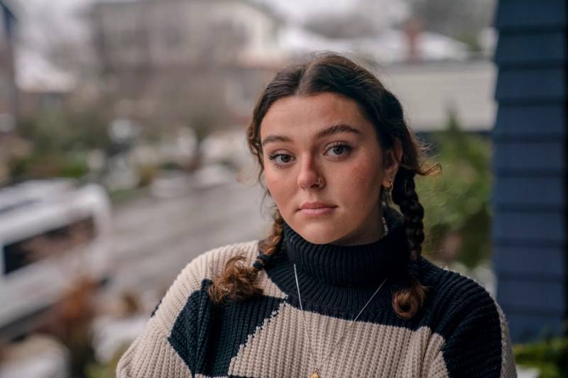 A young white woman with brown braided hair wearing a sweater looks at the camera.