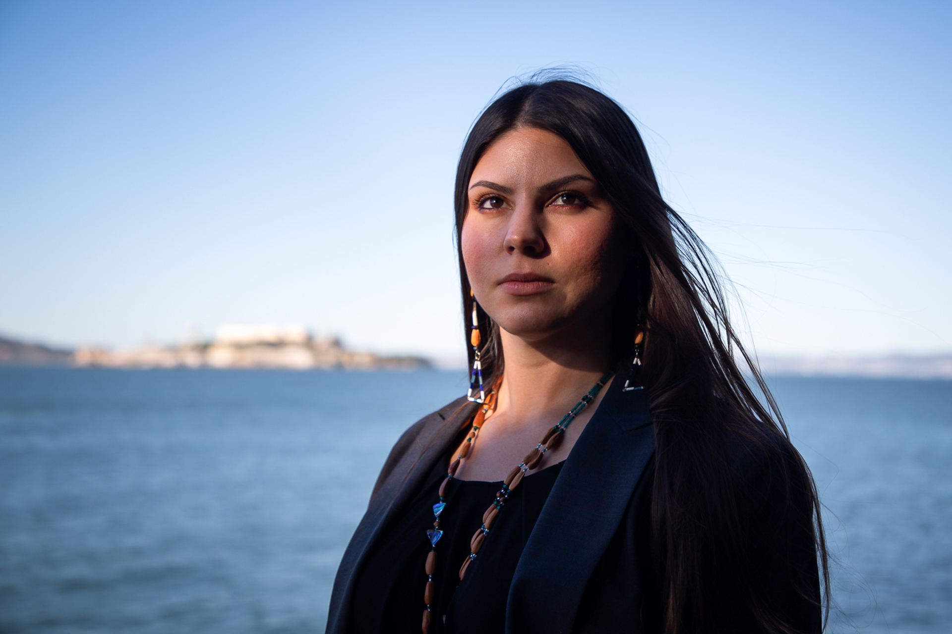 A Native American woman gazes into the camera. Her hair is long and dark, and she's wearing a beaded necklace and earrings. Behind her is the water, and just visible out of focus is Alcatraz Island.