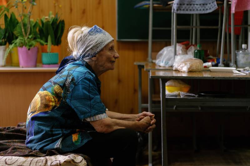 An elderly woman sits solemnly on her bed cot inside of a church office.