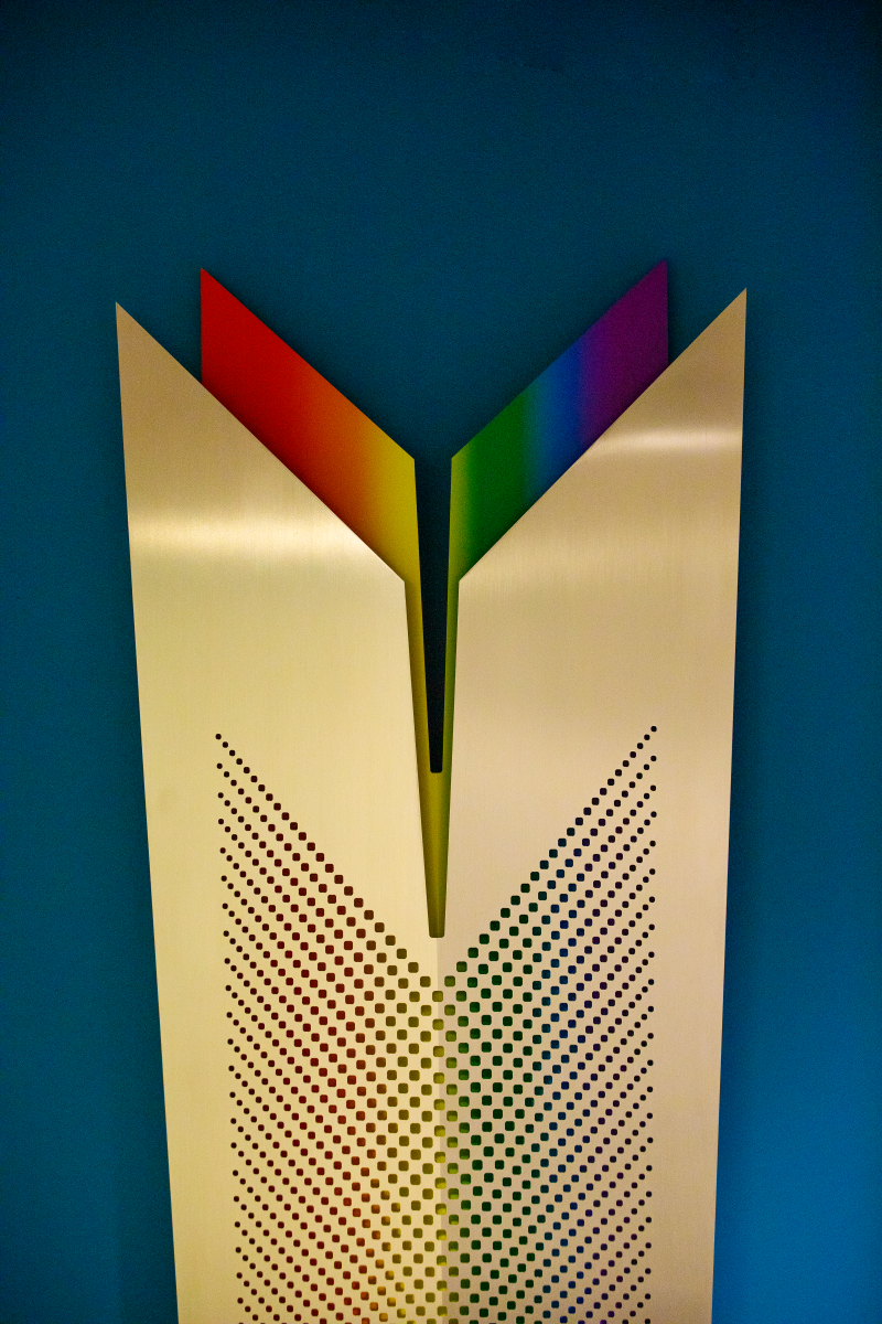 A gold and rainbow-colored metal sculpture hanging on a blue wall.