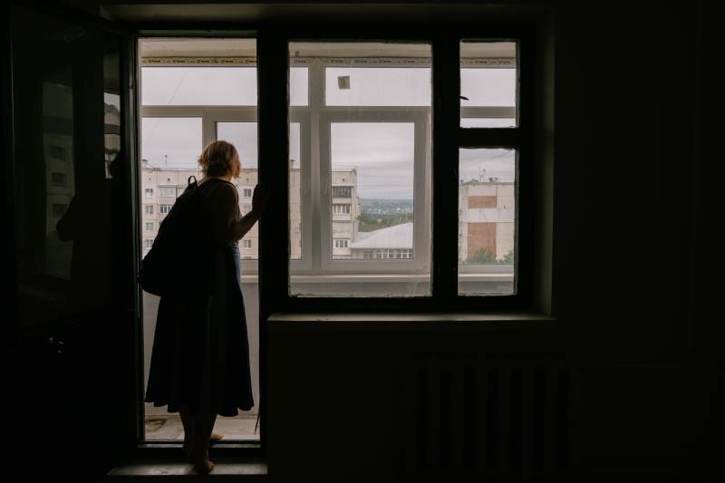 A woman stands framed in a doorway, which with adjacent windows frames and the wall are in shadow, to a terrace outside an apartment building.