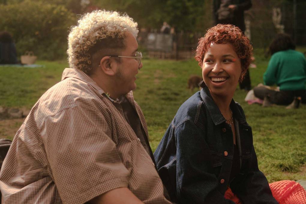 Two trans people of color sit in a park, with a hazy sunlight on their faces -- they look as if they're having a picnic, and are gazing happily at each other.