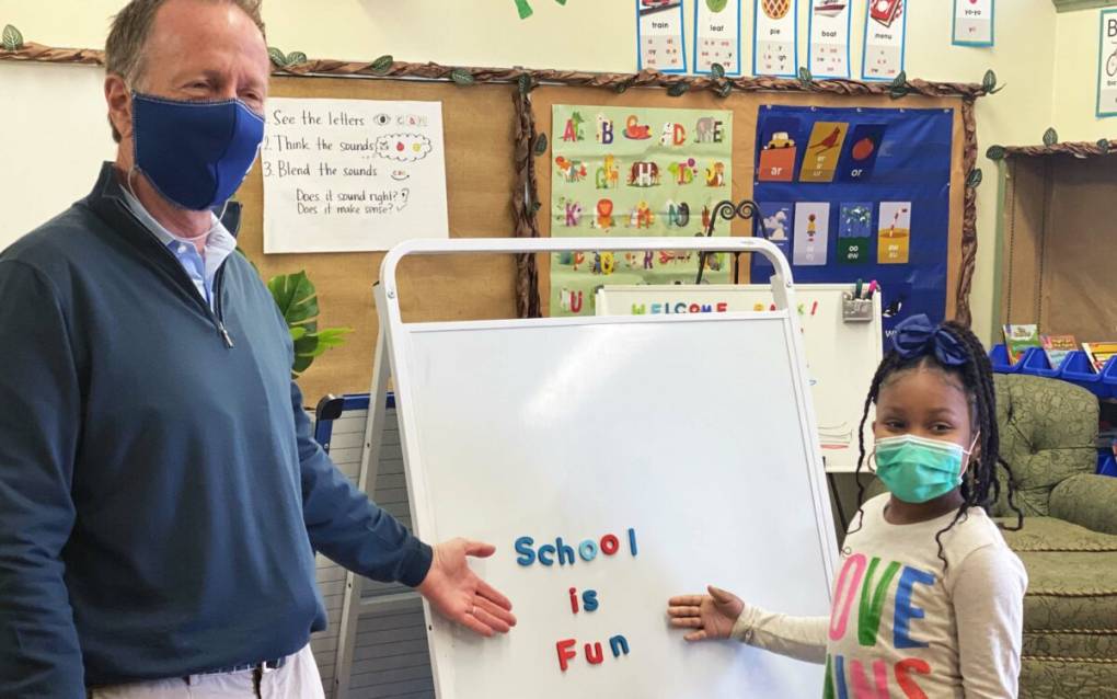 a tall white man and a young Black girl stand in a classroom in front of a white board where letters spell out 'school is fun'