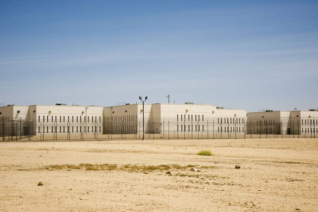 A white building behind high security fencing with desert in the foreground and a blue sky above.