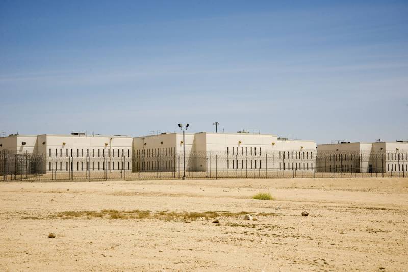 A white building behind high security fencing with desert in the foreground and a blue sky above.