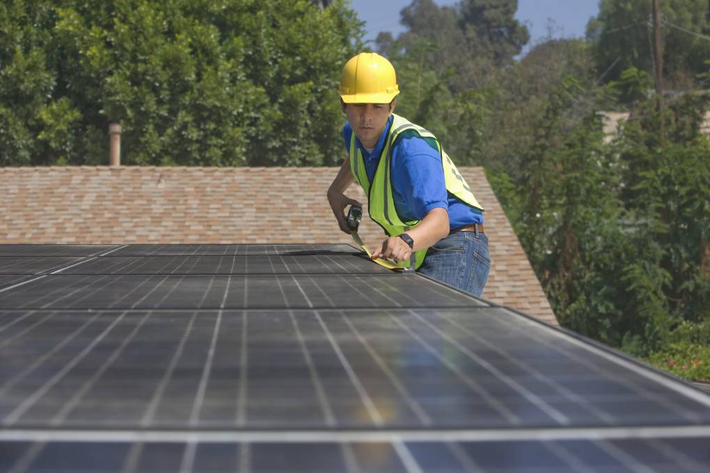 A rooftop solar panel worker with a yellow hard hat measures a solar panel on a roof with trees in the background.