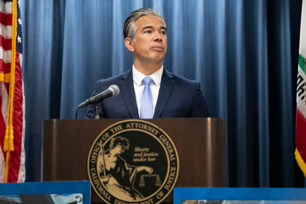 A Filipino man in a blue suit and tie speaks at a press conference behind a dais with the emblem of the office of the California attorney general on it.