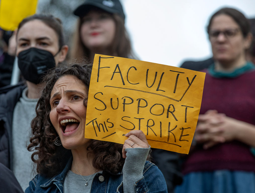 A woman in the foreground holds a sign that says "Faculty Support This Strike" with otherv faculty members around her. 