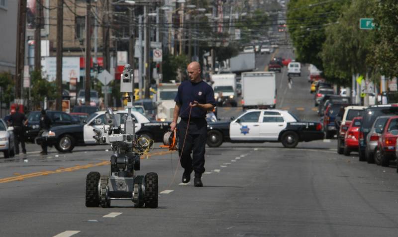 A man dressed in a police uniform holds a device that is attached to a robot in the middle of the street with police cars in the background.