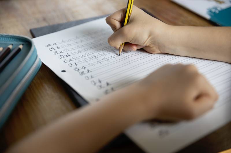 A child practices cursive handwriting in a notebook at a school desk.