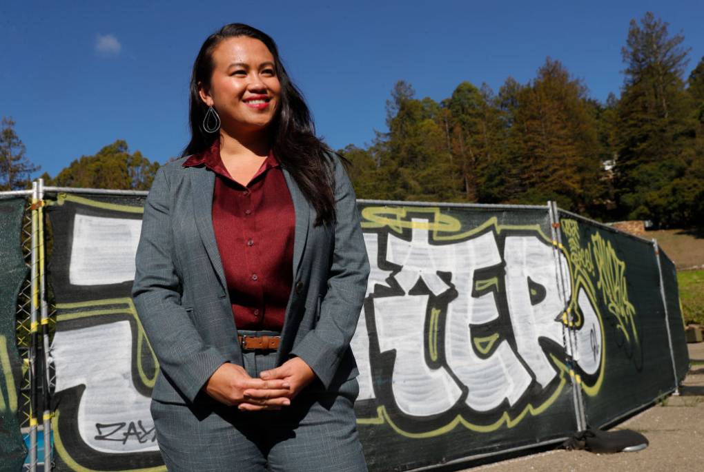 An Asian woman standing with her fingers crossed wearing a business suit with a red dress shirt outside behind a graffiti banner.