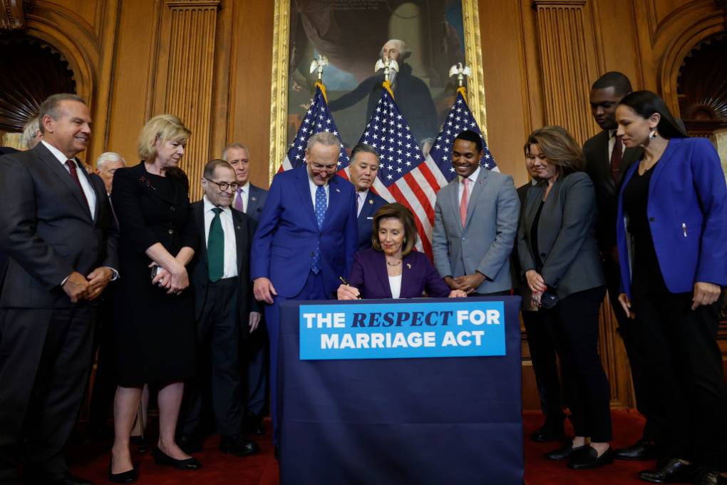A group of Senators and Representatives gather around a seated woman signing a bill at a dais with the words "The Respect for Marriage Act" written on it