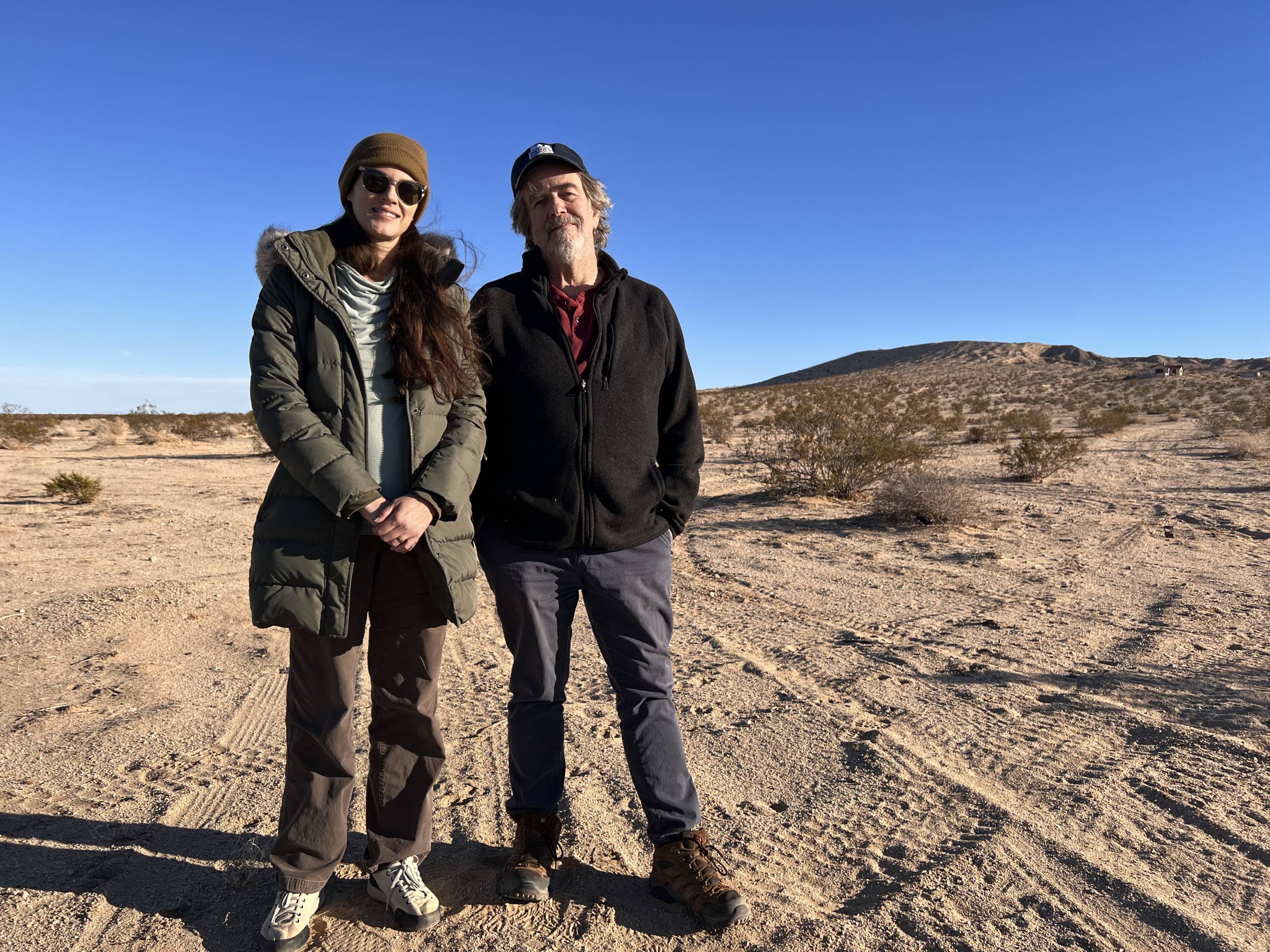 a woman and a man dressed warmly smile for a portrait in the desert against a blue sky