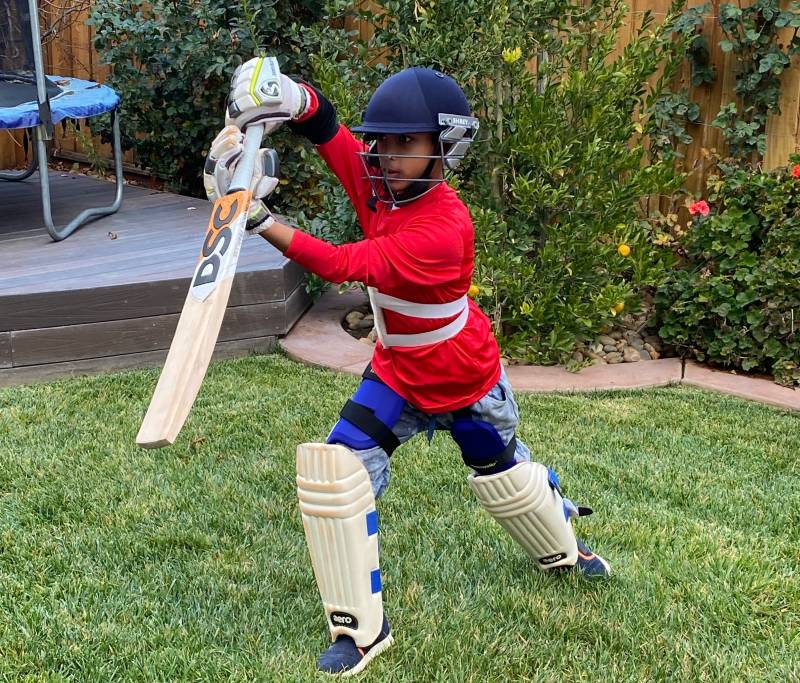 A young boy, dressed in cricket sports gear, holds a cricket bat upside down outdoors.