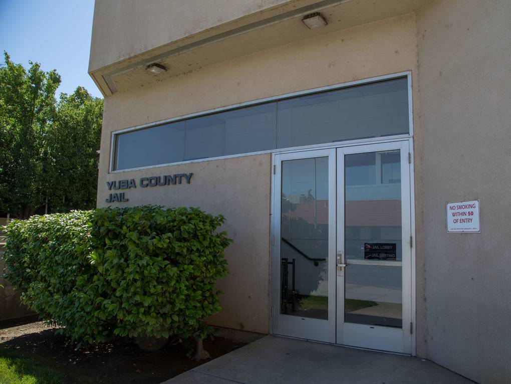 The outside of Yuba County Jail, the main entrance, a glass door in a drab old beige colored building with some bushes by the door.