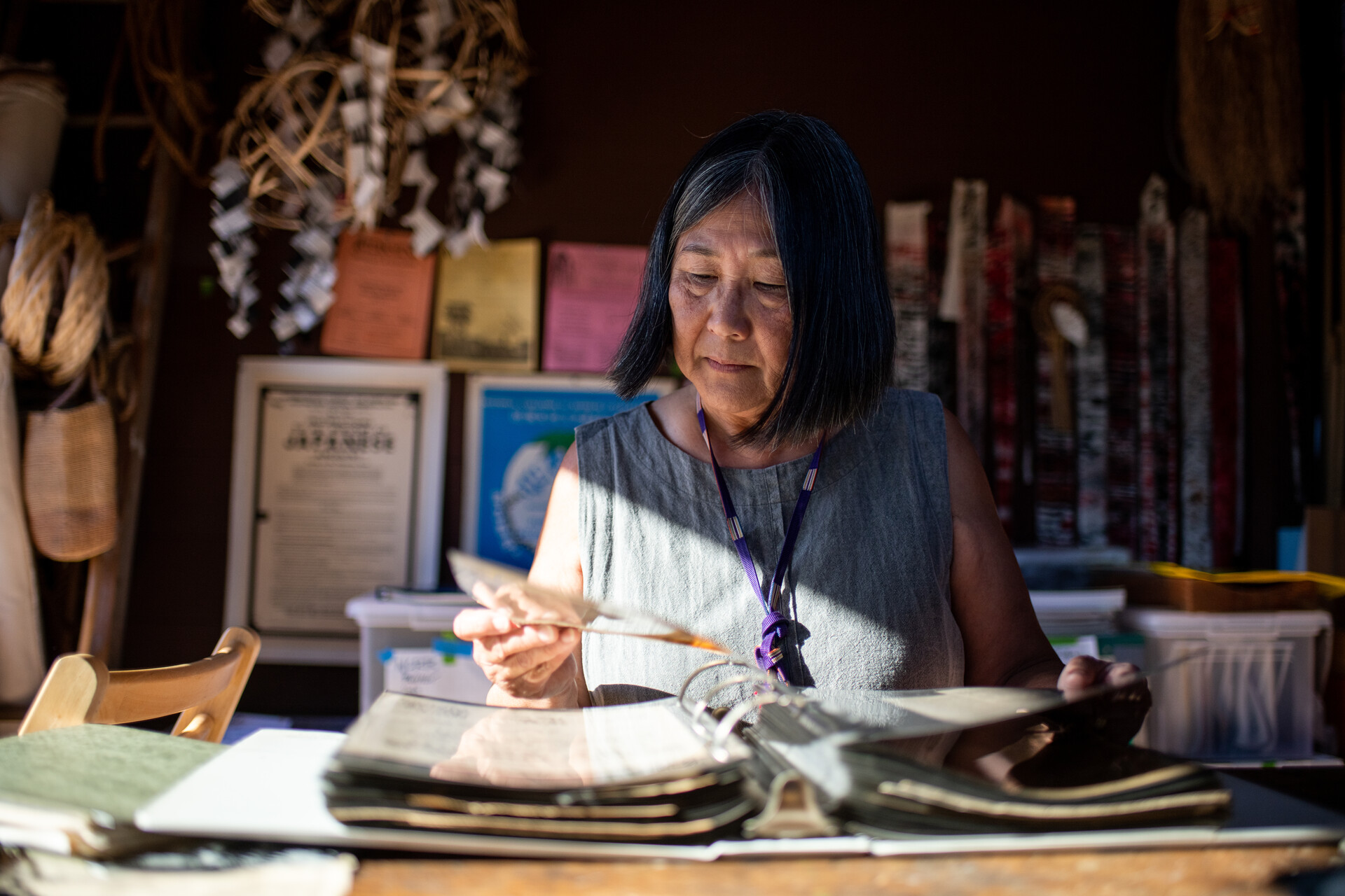 An older Asian woman with straight black hair with streaks of grey at the front wears a grey sleeveless shirt, and sits at a table surrounded by papers and books. She is holding one piece of paper in her right hand and looking at it.