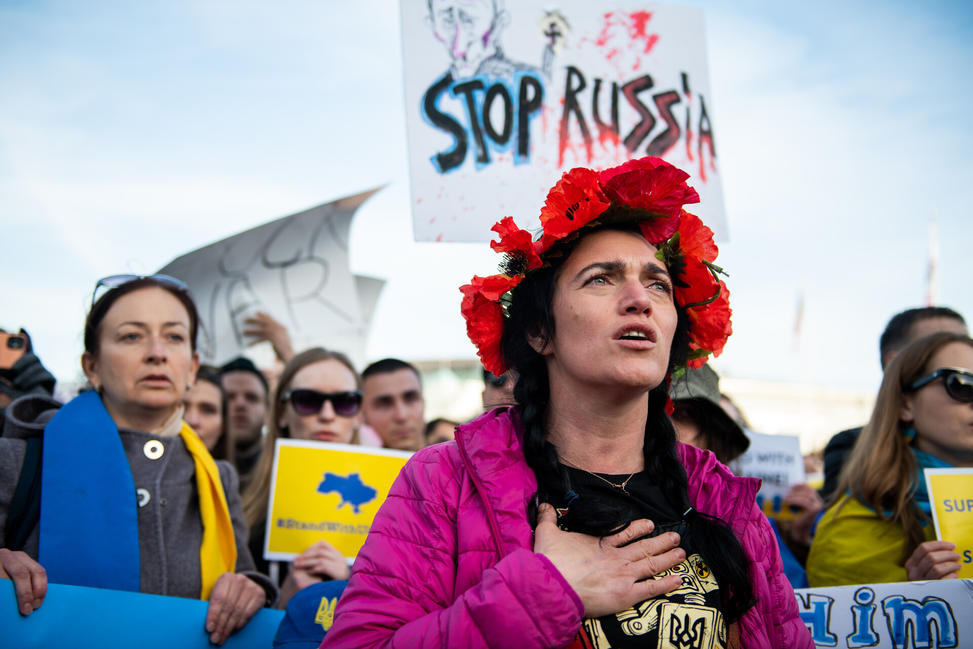 A woman with light skin stands in a crowd of protesters, wearing a pink jacket. A crown of red flowers is on top of her head, and she is holding her right hand to her chest and appears to be singing. A sign saying 