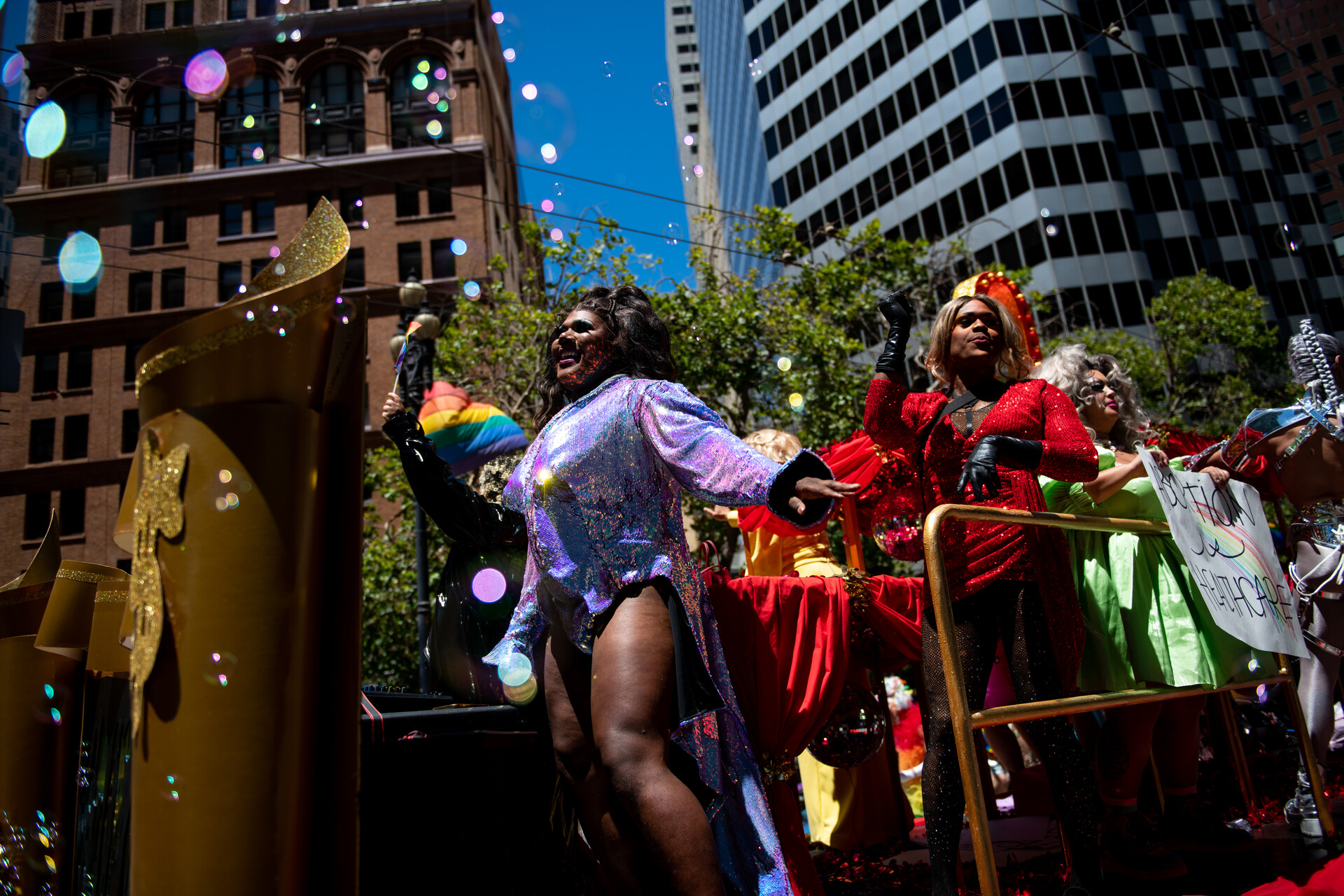 Drag queens dance on a float at San Francisco Pride with tall building and a bright blue sky behind them. At the front is Nicki Jizz, a Black drag queen wearing a short iridescent purple dress.