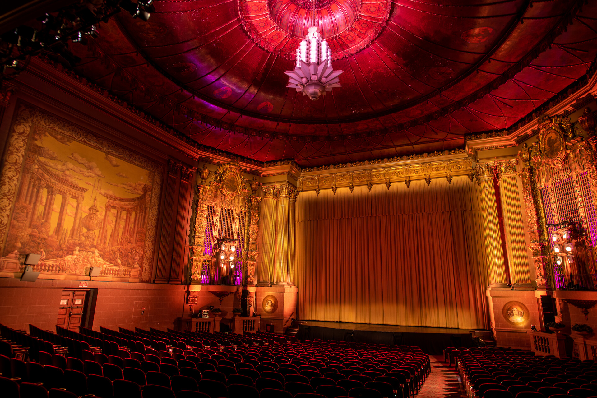 An ornate theater interior in the Art Deco style, bathed in warm tones of red and gold. The photograph is taken from the back of the theater, facing the stage and its gold draped curtain.