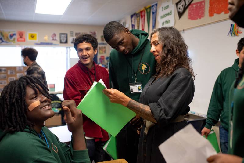 A woman with light-brown skin, a tattoo sleeve on her left arm, and long gray-brown hair appears to read out loud from a piece of paper she holds as a Black male student, a Latinx male student and a Black female student stand or sit near her inside a high school classroom. The mood is relaxed and happy.