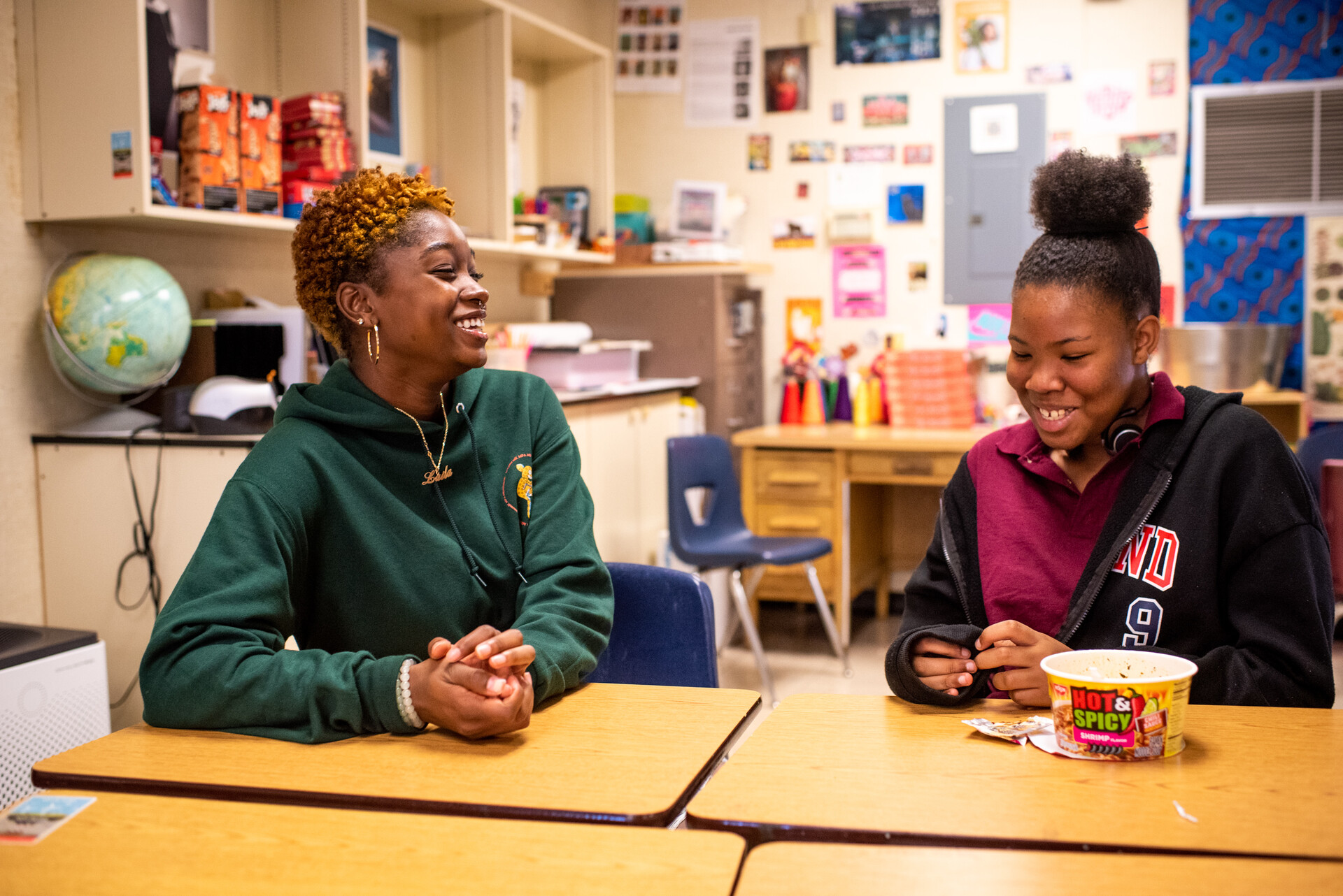 Two black female high school students sit in class and laugh together.