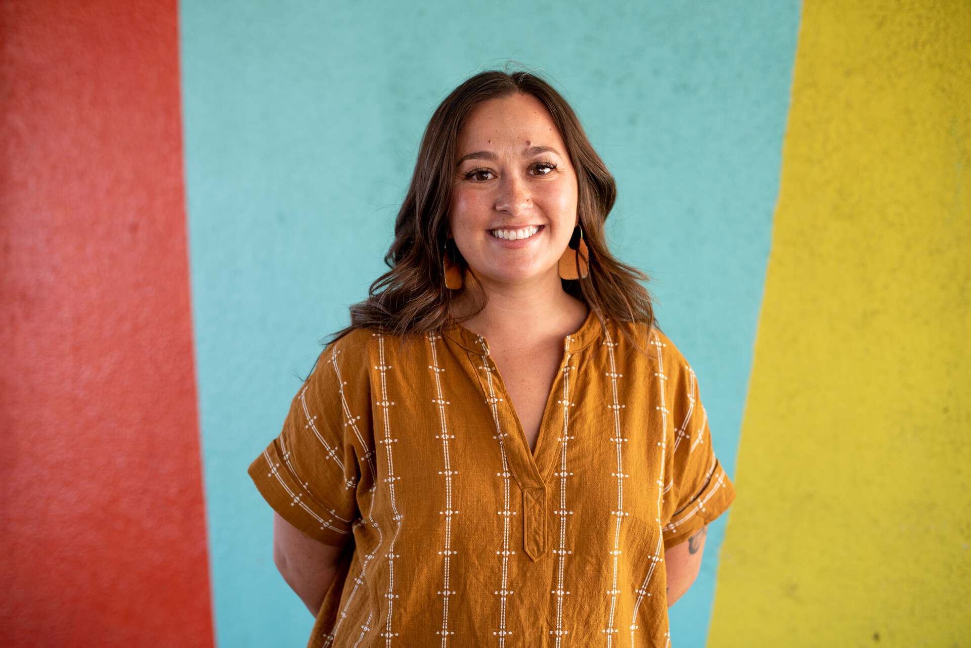 A Latina woman with long brown hair smiles at the camera with a colorful wall in the background.