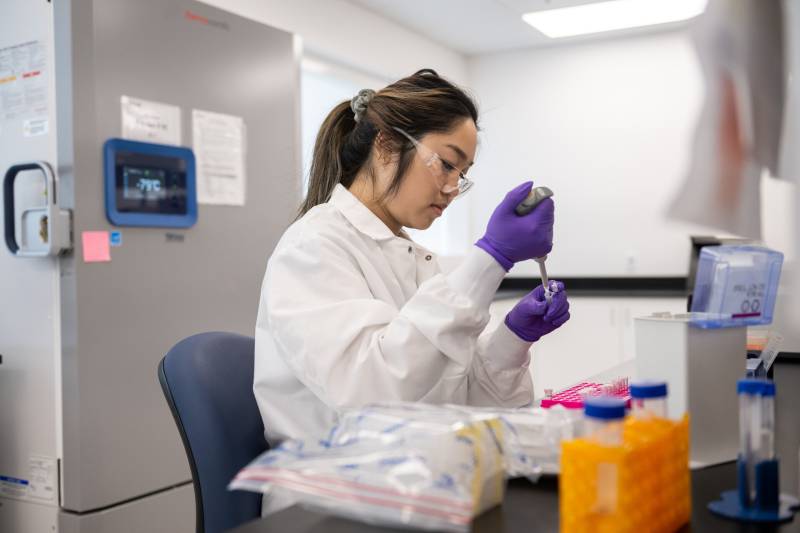 a woman who appears to be of Asian descent works in a lab wearing a white coat and purple gloves