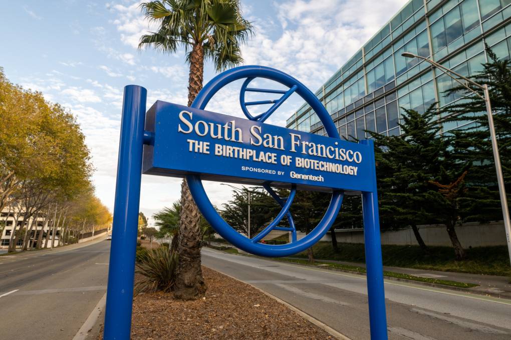A blue sign for South San Francisco, 'The Birthplace of Biotechnology,' against a blue sky and a palm tree with an office building in the background