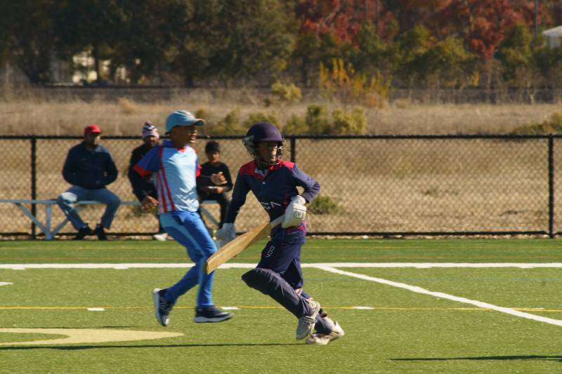 A young boy starts to run while holding a cricket bat with people in the background.