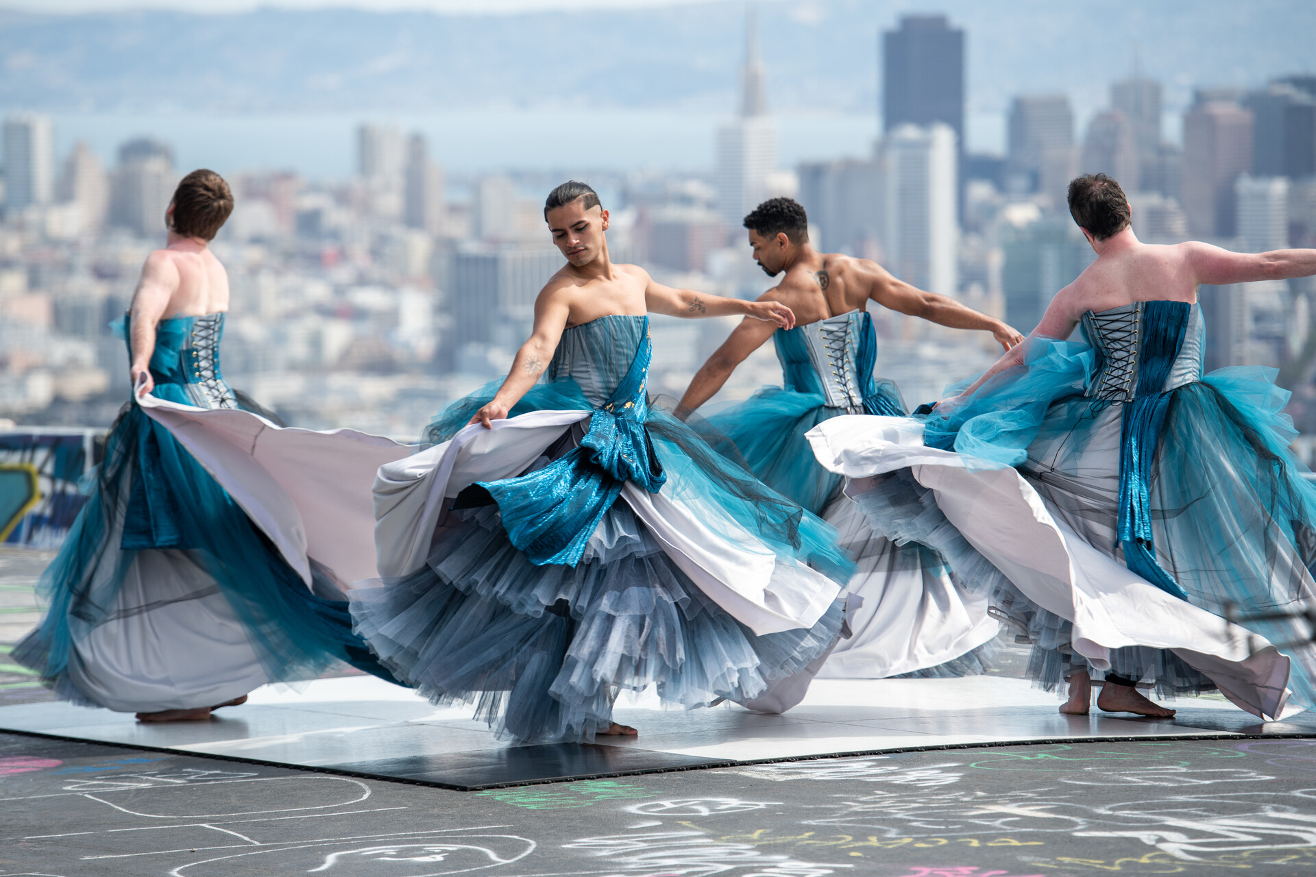 A group of dancers twirl dressed in strapless dresses that are aqua blue and white, photographed mid-twirl. Behind them is a striking view of the downtown San Francisco skyline.