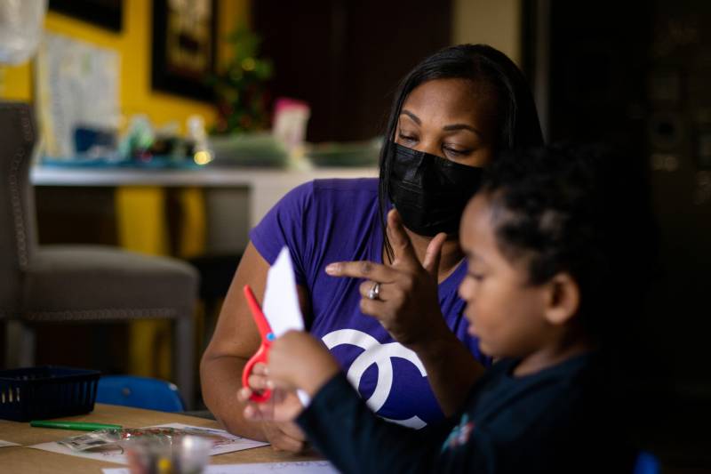A woman wearing a purple t-shirt and black face mask sits next to a child holding red scissors.