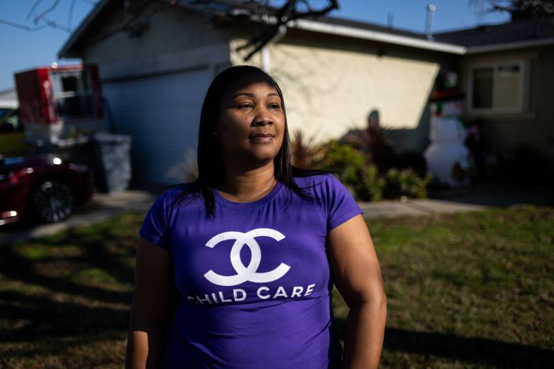 A Black woman wearing a purple t-shirt that says "Child care" stands outside.