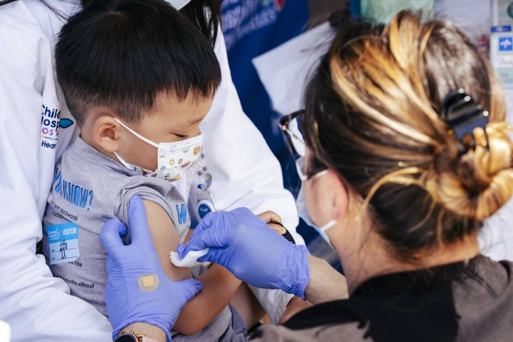 A small Asian child wearing a spotted face mask sits on the lap of an unseen person wearing a white coat, while a medical professional wearing blue surgical gloves leans over them and swabs their arm.