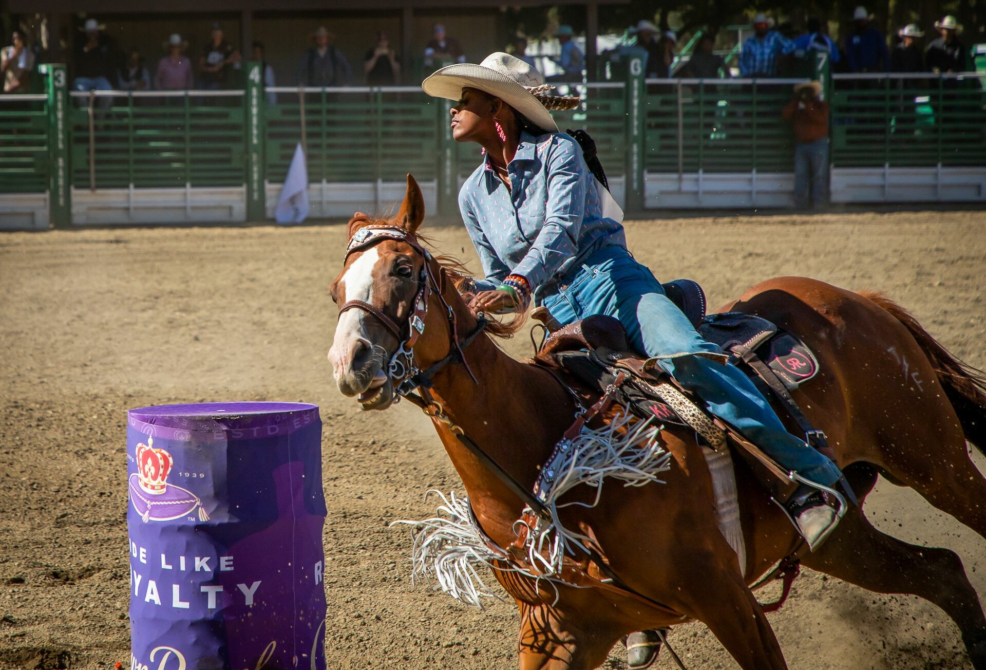 A Black woman wearing double denim rides a tan horse in a sandy arena watched by a crowd. They are photographed leaning forward and to the side as both horse and rider pivot around a purple painted drum.