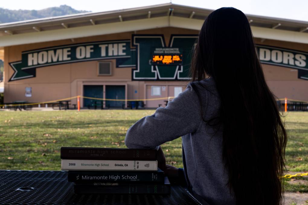 a young woman with long dark hair sits at a table with books, with her back to the camera, facing a school that has a sign that reads 'home of the matadors'