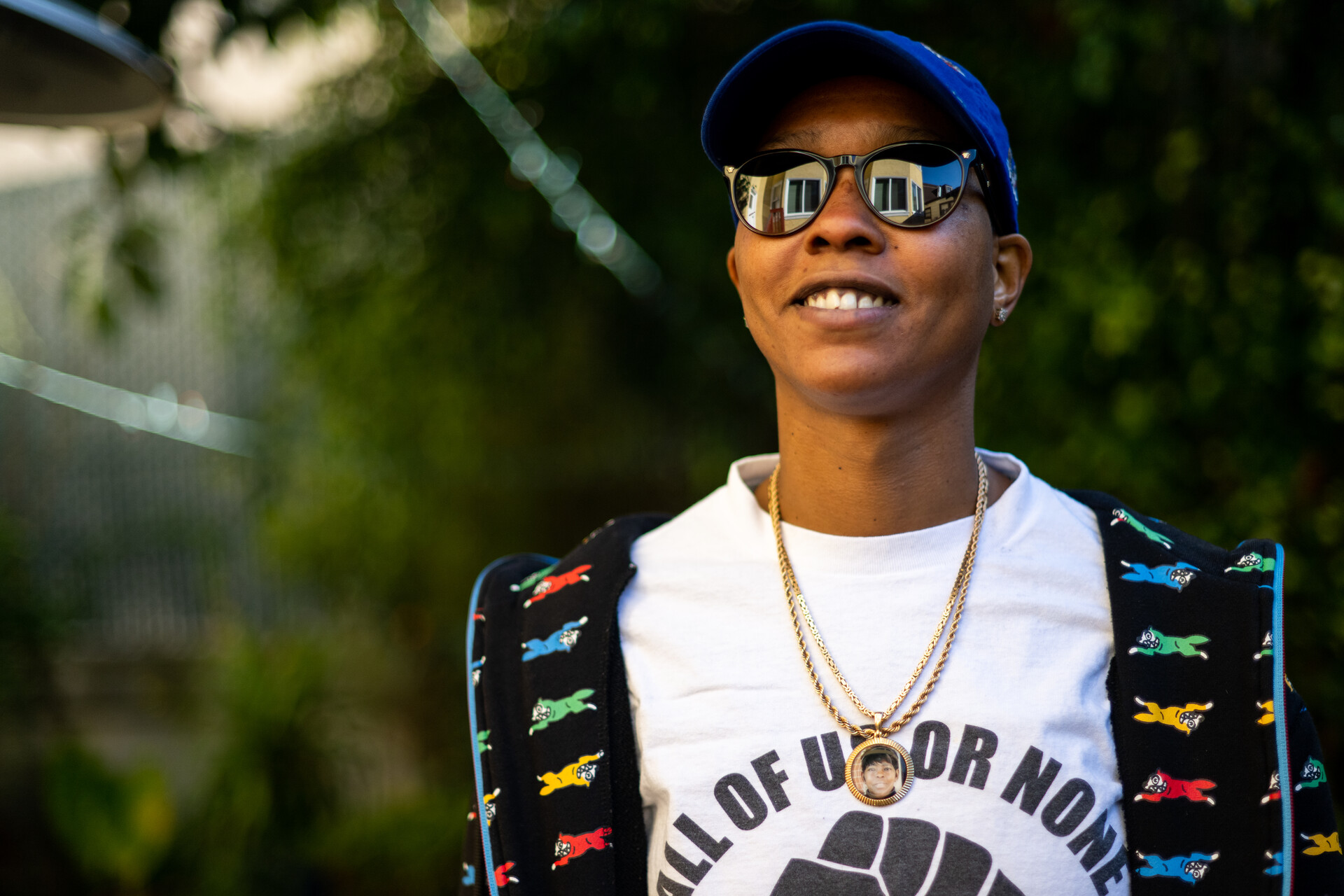 A Black woman with a white shirt that reads "All of Us or None" and sunglasses with her house reflected in the lenses, wearing a blue cap and smiling.