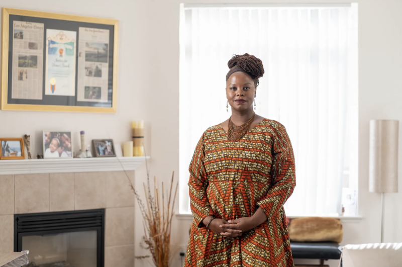 A Black woman wearing a dress with an orange and brown design pattern stands in a house with her fingers interlocked.
