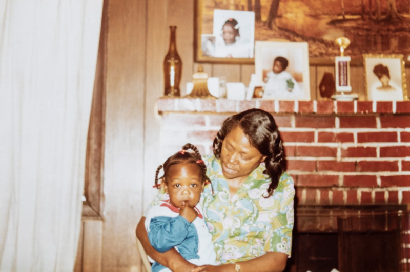 A vintage photo of a Black woman wearing a floral printed shirt holds a young Black girl in a house.
