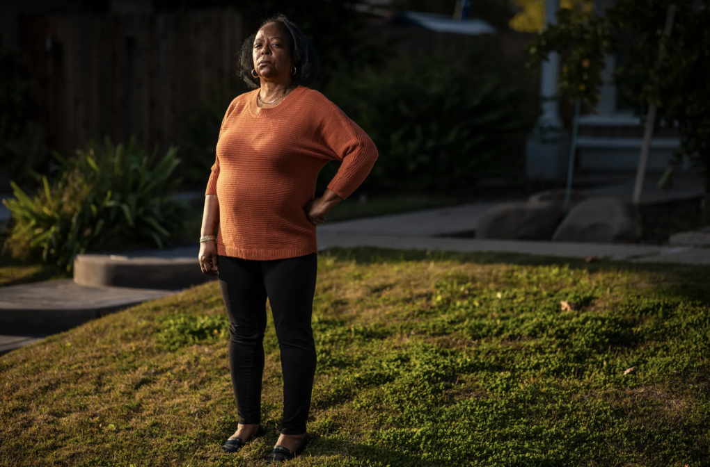 A Black woman wearing an orange top and black pants stands outside with her hand on her hip.