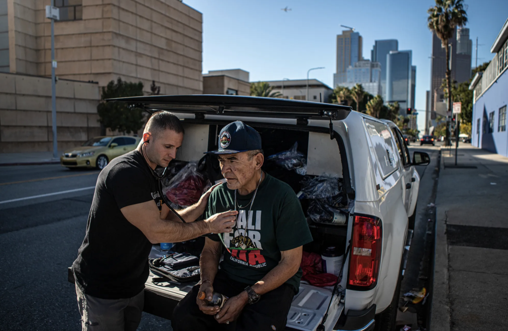 A man wearing a black t-shirt places the receiving end of a stethoscope on a man's chest who is wearing a California Republic hat and t-shirt and sitting on a pullout trunk of a car.