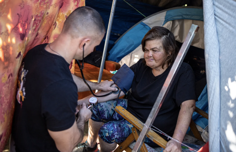 A man wearing a black t-shirt uses a stethoscope on a woman's arm in a tent.