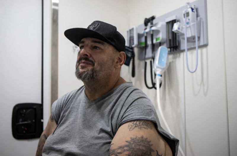 A man with a black Raiders hat and gray shirt sits down in a medical facility.