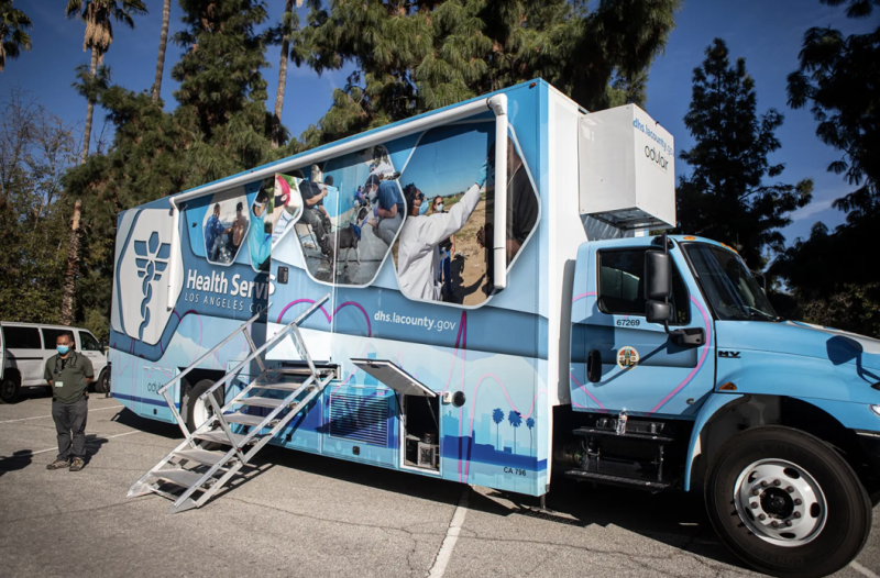 A light blue medical truck with several photos of people and a medical logo on the side with a ramp and stairs leading inside.