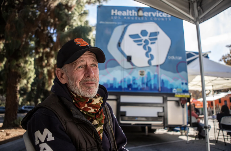 A man wearing black hat, camouflage scarf and black jacket outside near a truck with a medical logo.