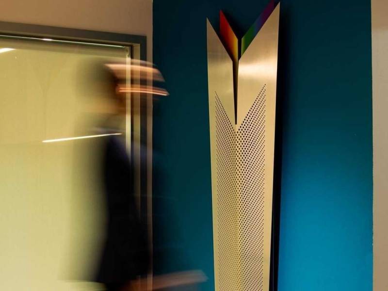 a blurry figure approaches a metal shield-like sculpture which is mounted on a blue wall