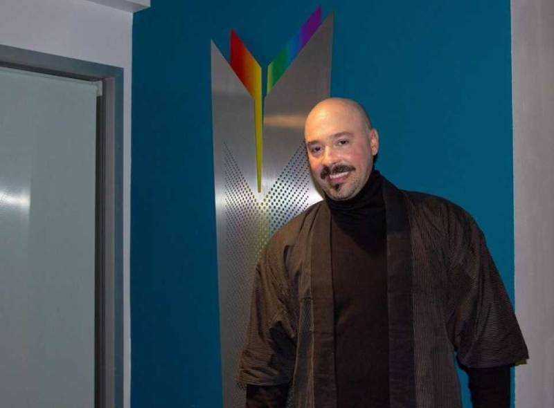a young-looking, bald Latino man with a dark brown mustache and goatee, wearing a brown shirt and coat, smiles in front of a metal and rainbow-hued sculpture hanging on a blue interior wall. The sculpture is roughly a couple feet wide and 6 inches taller than a nearby doorframe.