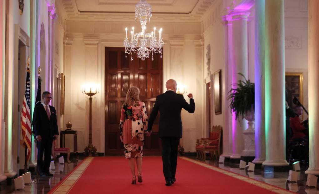 President Joe Biden and First Lady Jill Biden, seen from behind, walk through an ornate hallway lit with rainbow colors.