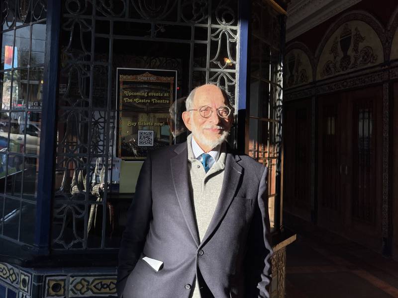 an older man in a dark blue suit with a white beard and glasses smiles in front of a theater