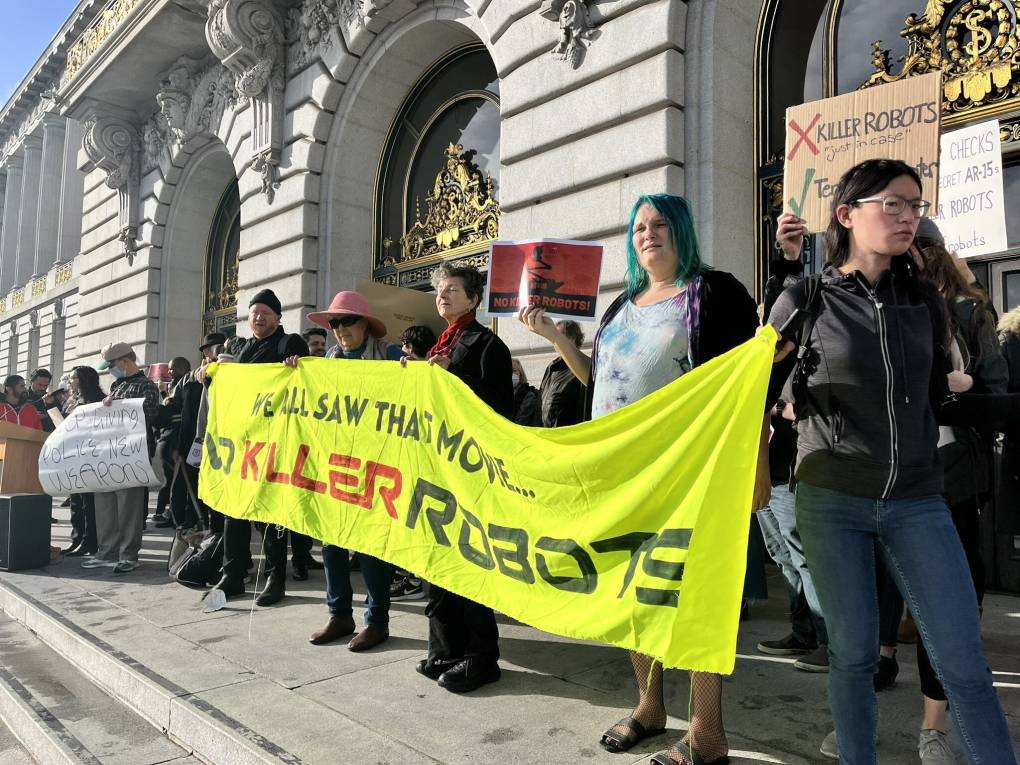 Several people stand outside a building holding a neon green sign that says "We all saw that movie..no killer robots."